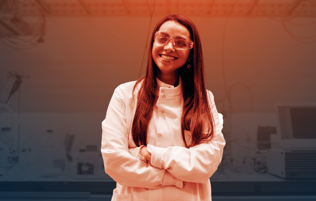 A latinx woman in lab coat and protective glasses with arms crossed with laboratory in background.